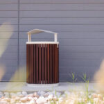 A brown and white outdoor trash can stands on a concrete surface in front of a gray paneled wall, with rocks and grass in the foreground.