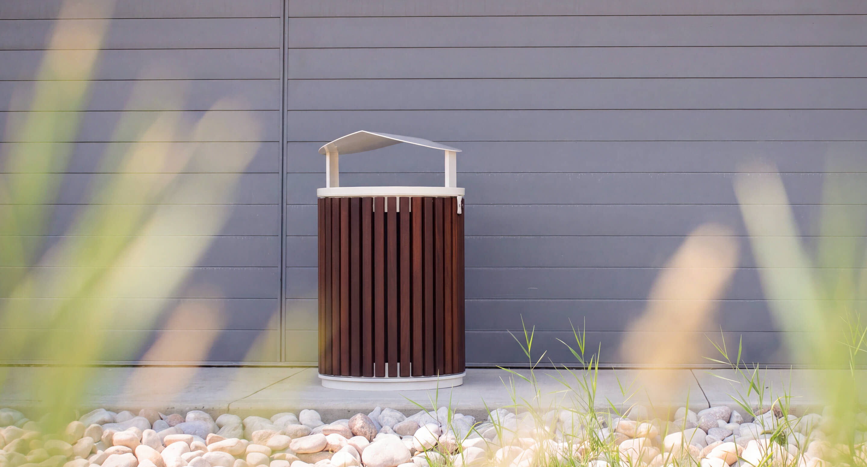 maglin_mtr-0720-trash-container_carousel A brown and white outdoor trash can stands on a concrete surface in front of a gray paneled wall, with rocks and grass in the foreground.