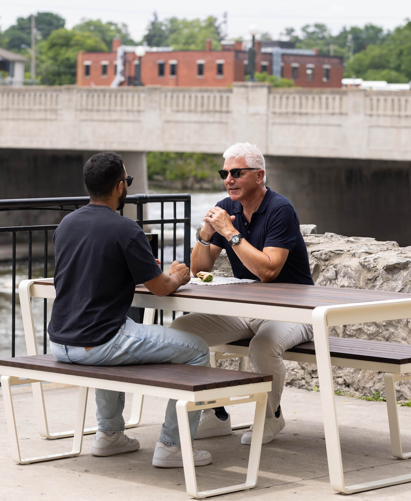 Two men sit across from each other at an outdoor table near a stone wall, talking. A bridge and buildings are visible in the background.