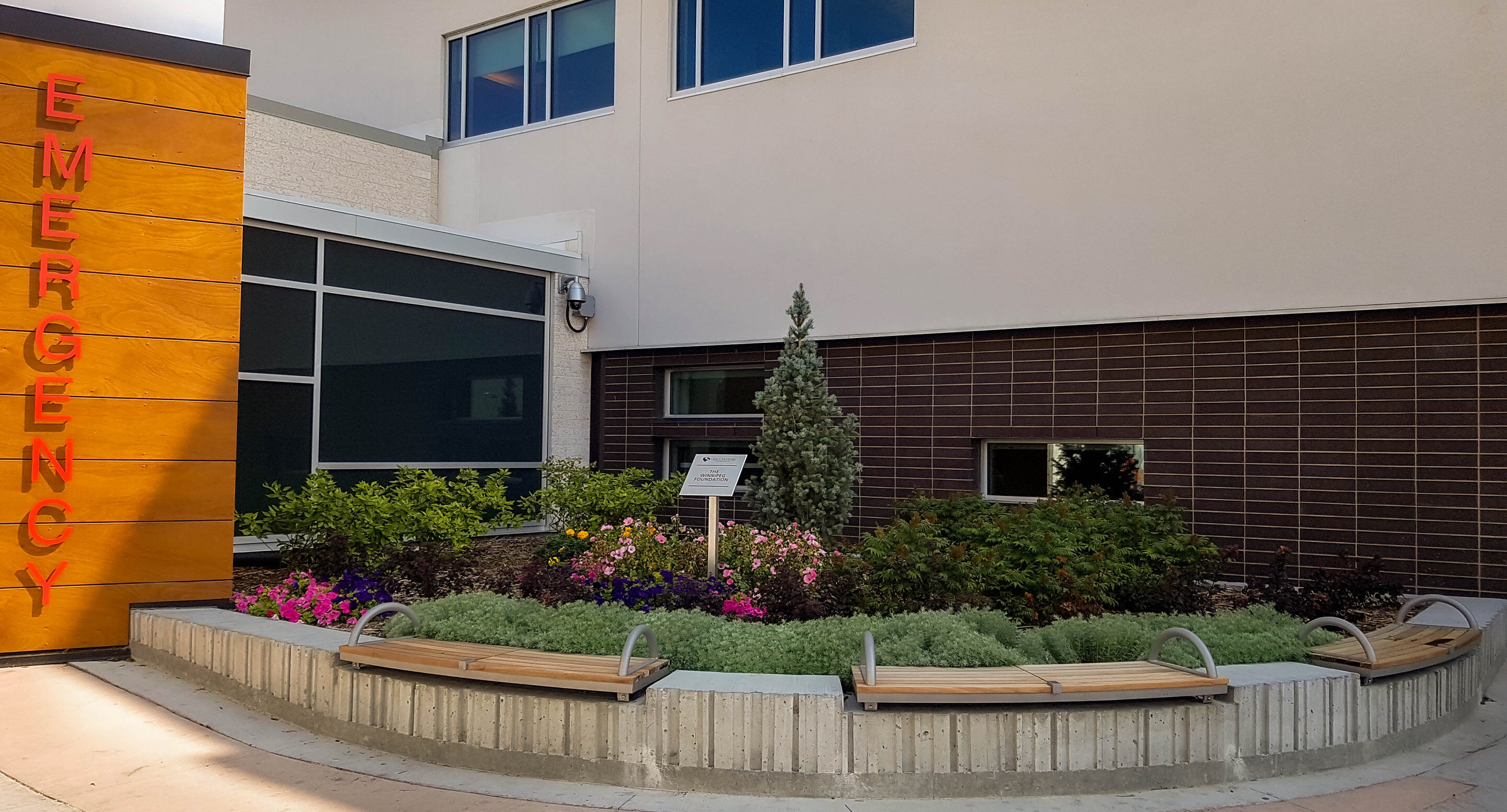 grace-hospital-emergency-department-ogden-wall-mount-1 Entrée des urgences, avec trois bancs en bois incurvés placés devant un parterre paysager adossé à la façade du bâtiment.