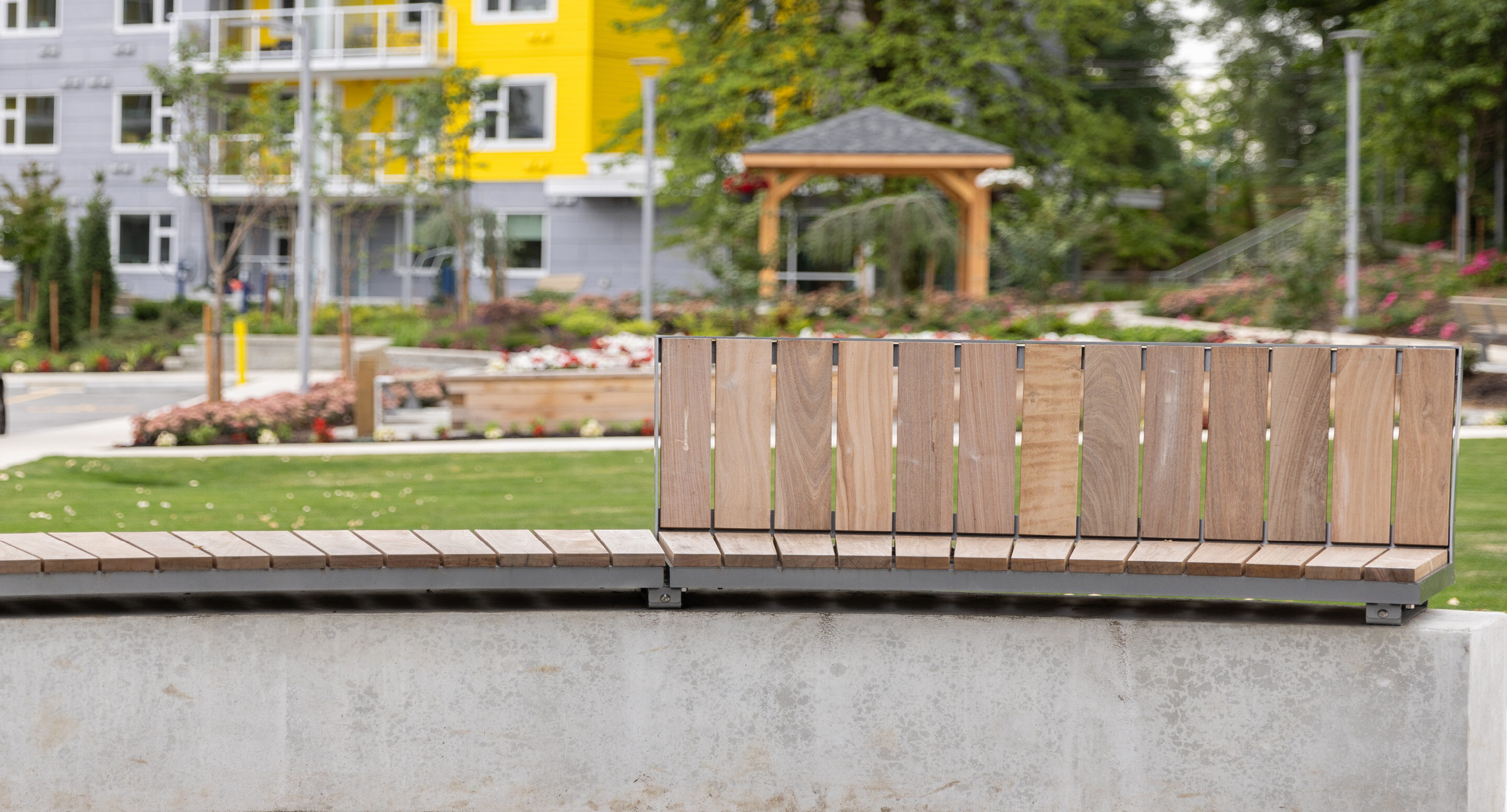 granly-gardens-dania-society-ogden-close Un banc en lattes de bois installé contre un mur en béton dans un parc.