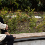 Une personne aux longs cheveux blonds est assise sur un banc en bois incurvé dans un jardin.