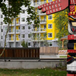 A colorful wooden totem pole stands in the foreground, with a curved wooden bench and the Granly Gardens apartment building in the background.