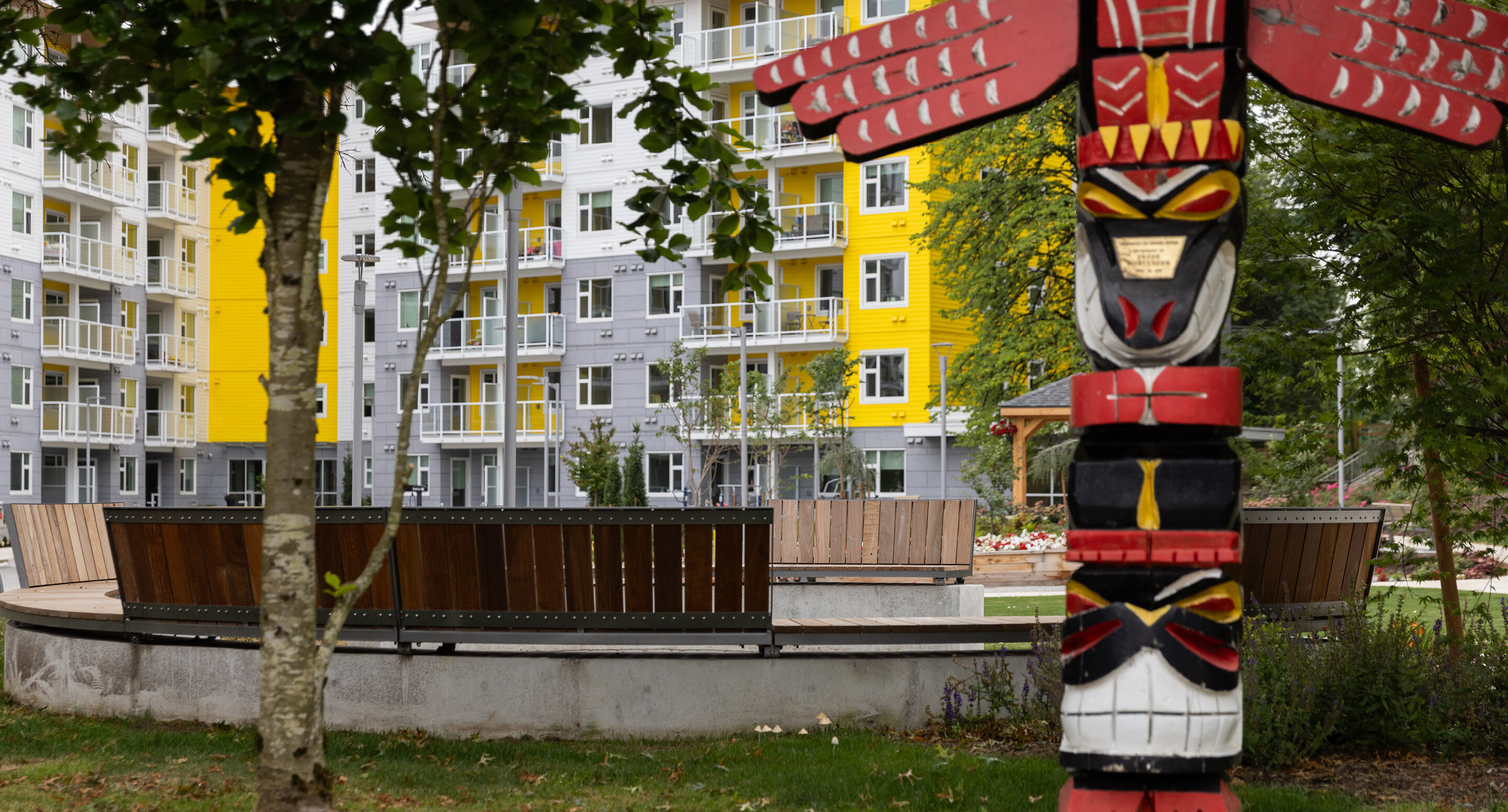 granly-gardens-dania-society-ogden-totem-pole Un totem sculpté et peint se dresse dans un parc, près d'un banc en bois incurvé et d'un immeuble jaune.