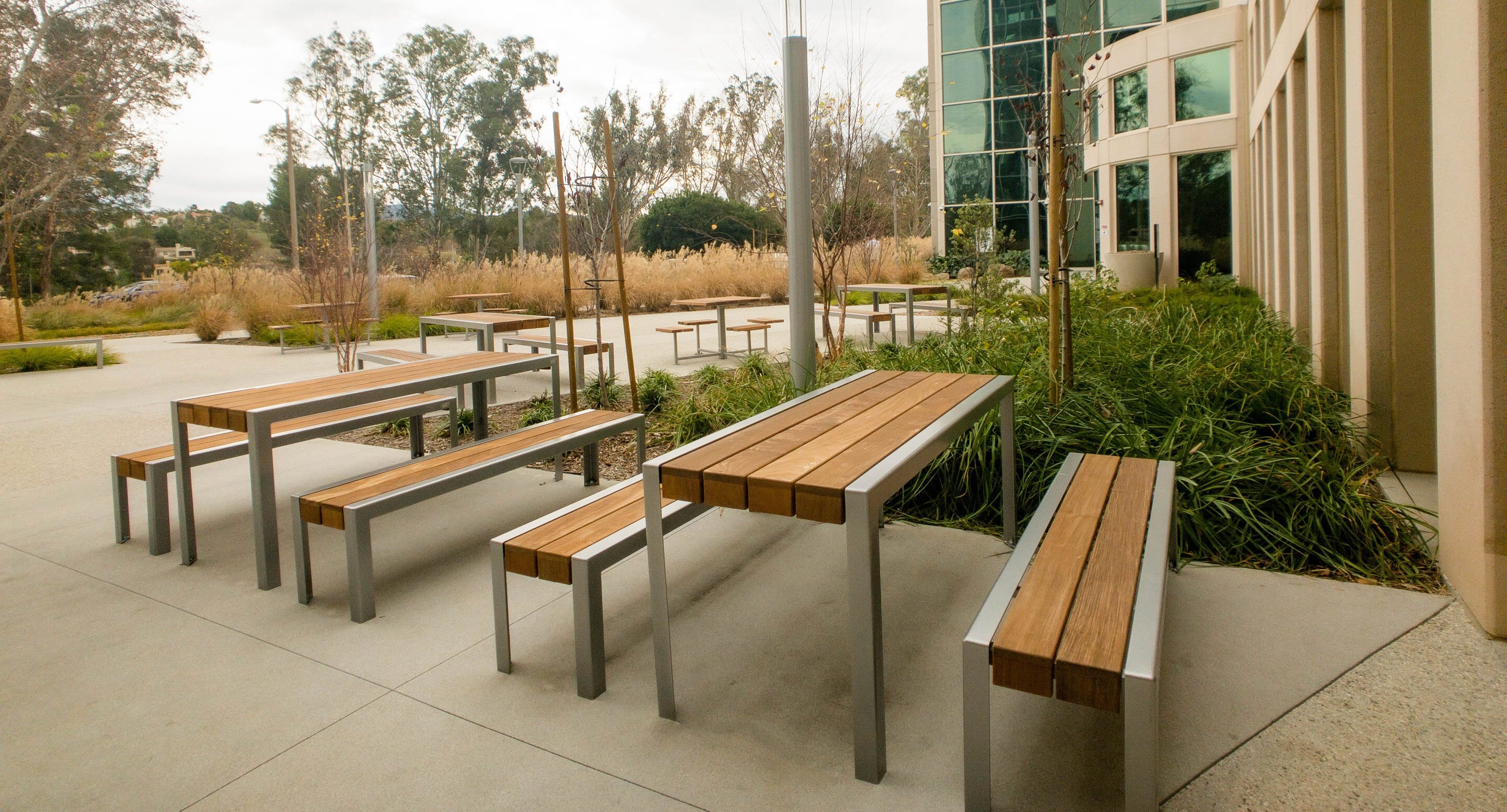 kaiser-permanente-santa-clarita-1050-benches-tables-400-cluster Terrasse extérieure équipée de plusieurs tables et bancs en bois, aménagée sur une esplanade en béton à côté d'un bâtiment aux façades vitrées.