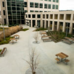 An outdoor courtyard with multiple wooden picnic tables, a large Kaiser Permanente office building with glass windows, and a “welcome” sign.