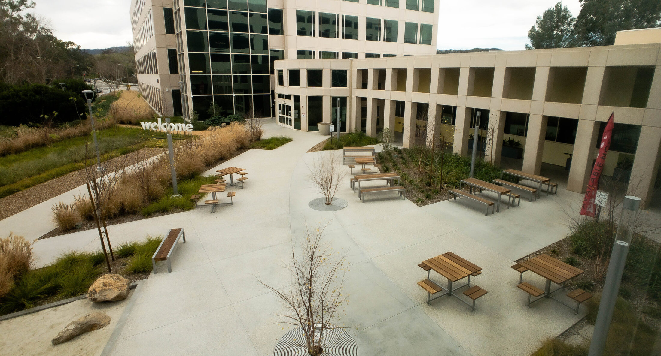 An outdoor courtyard with multiple wooden picnic tables, a large Kaiser Permanente office building with glass windows, and a “welcome” sign.