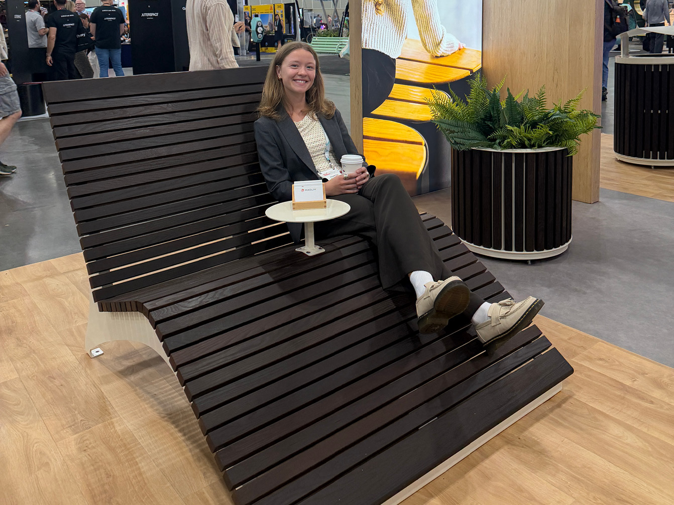 A woman holds a coffee cup while sitting on a Maglin wooden lounge chair with an integrated side table at the ASLA 2026 trade show.
