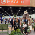 A group of eleven people posing for a photo in front of a Maglin trade show booth at ASLA 2026, featuring a wooden eagle sculpture and green metal benches.