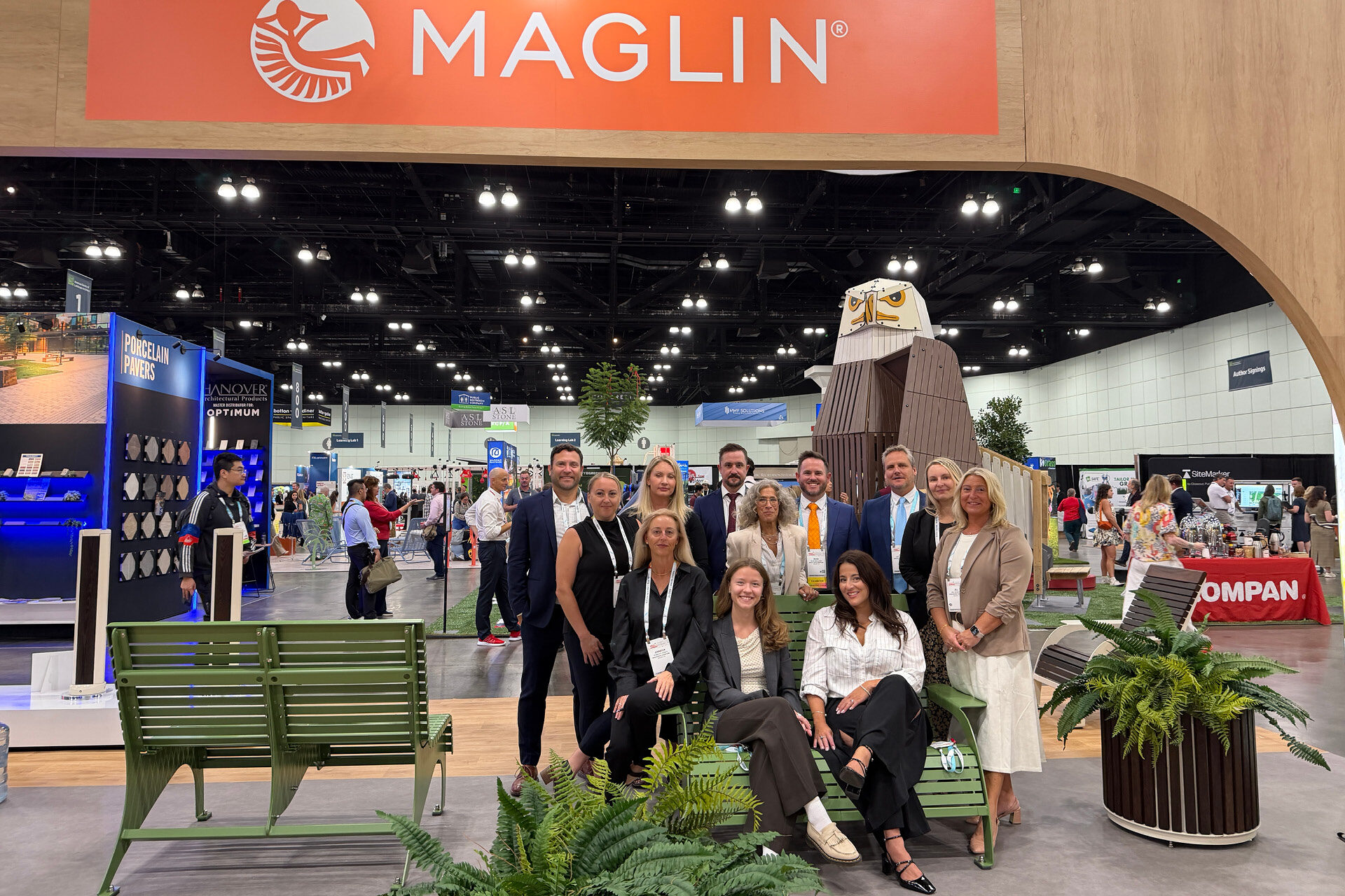 A group of eleven people posing for a photo in front of a Maglin trade show booth at ASLA 2026, featuring a wooden eagle sculpture and green metal benches.