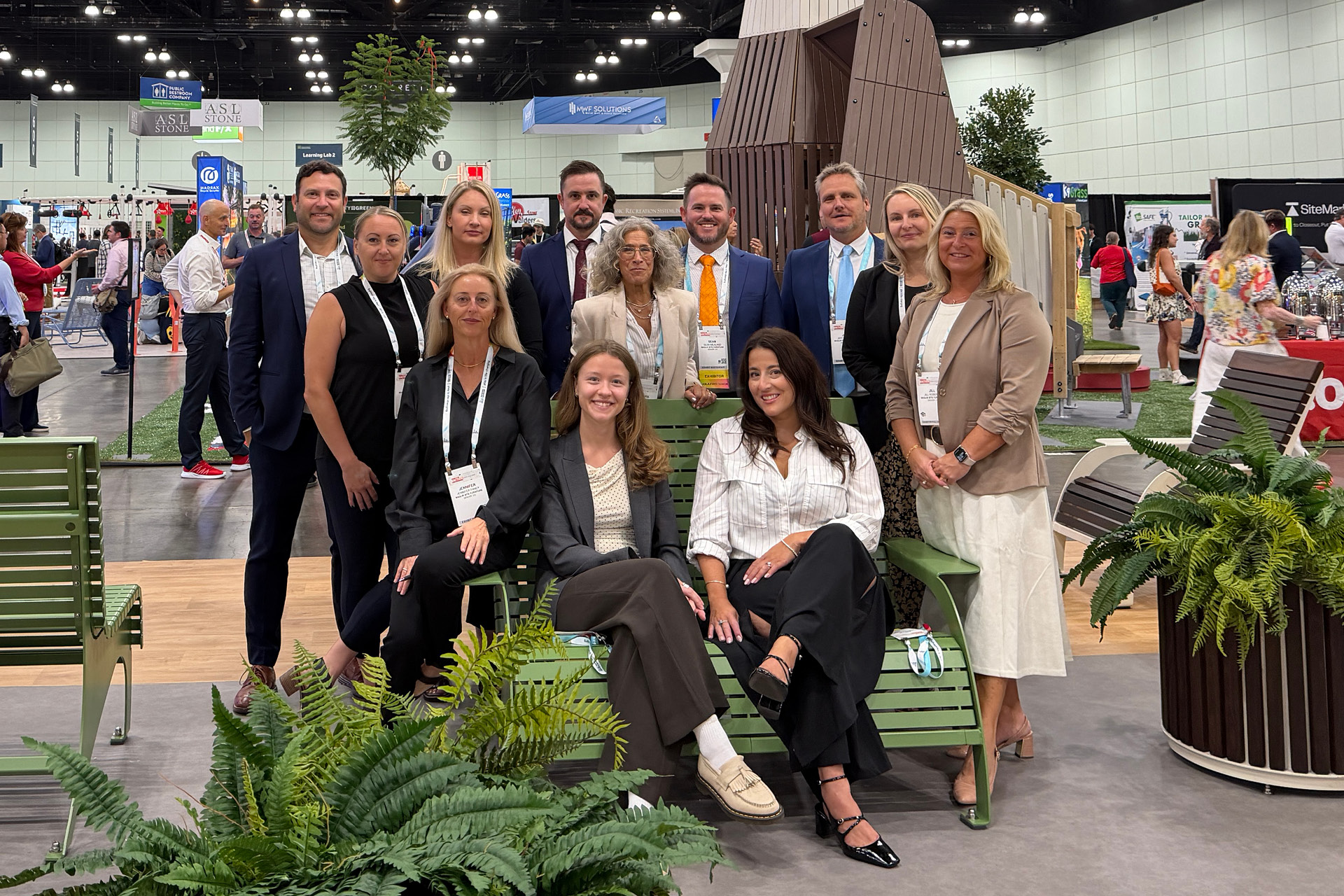 Eleven professionals pose for a photo at the Maglin booth during the ASLA 2026 trade show, with some seated on a green metal bench and others standing behind them.