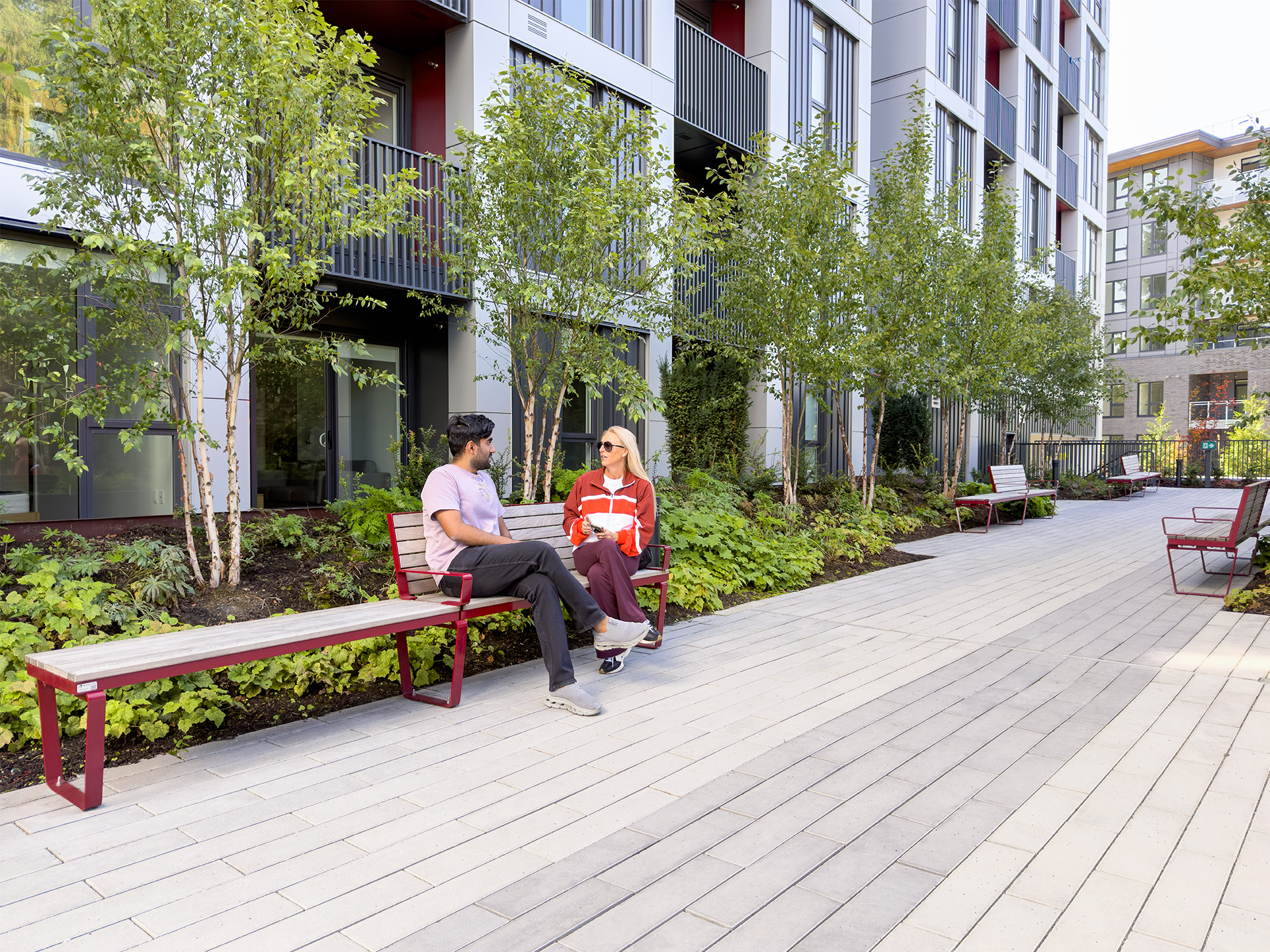 A man and woman sitting on a Maglin metal-framed Ogden bench on a paved walkway outside a modern apartment building.