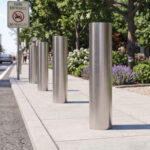 A row of cylindrical metal bollards lining a park entrance sidewalk next to a road.