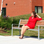 Une femme vêtue d'une robe rouge est assise sur un banc de parc devant un immeuble en briques.