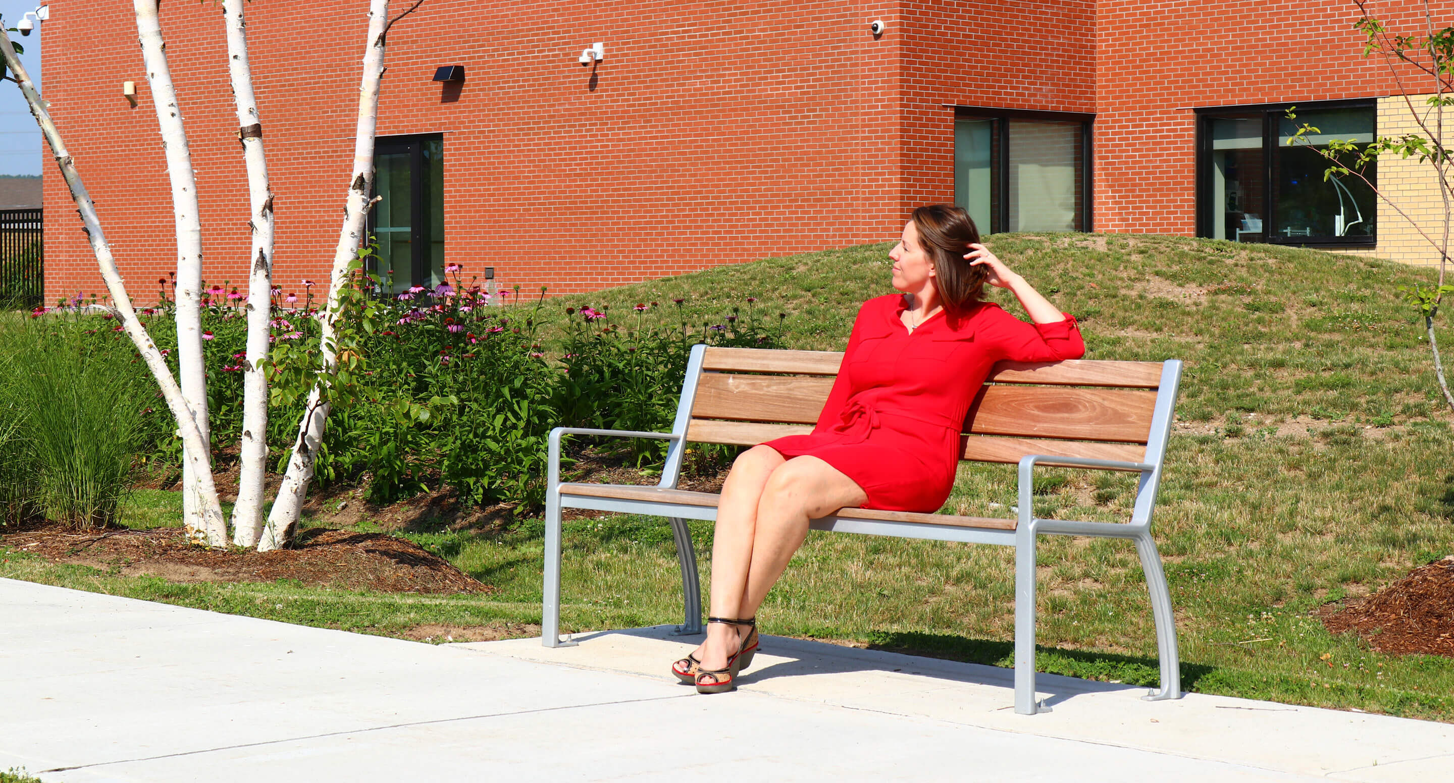 milton-district-hospital-970-bench-kyra-sitting Une femme vêtue d'une robe rouge est assise sur un banc de parc devant un immeuble en briques.