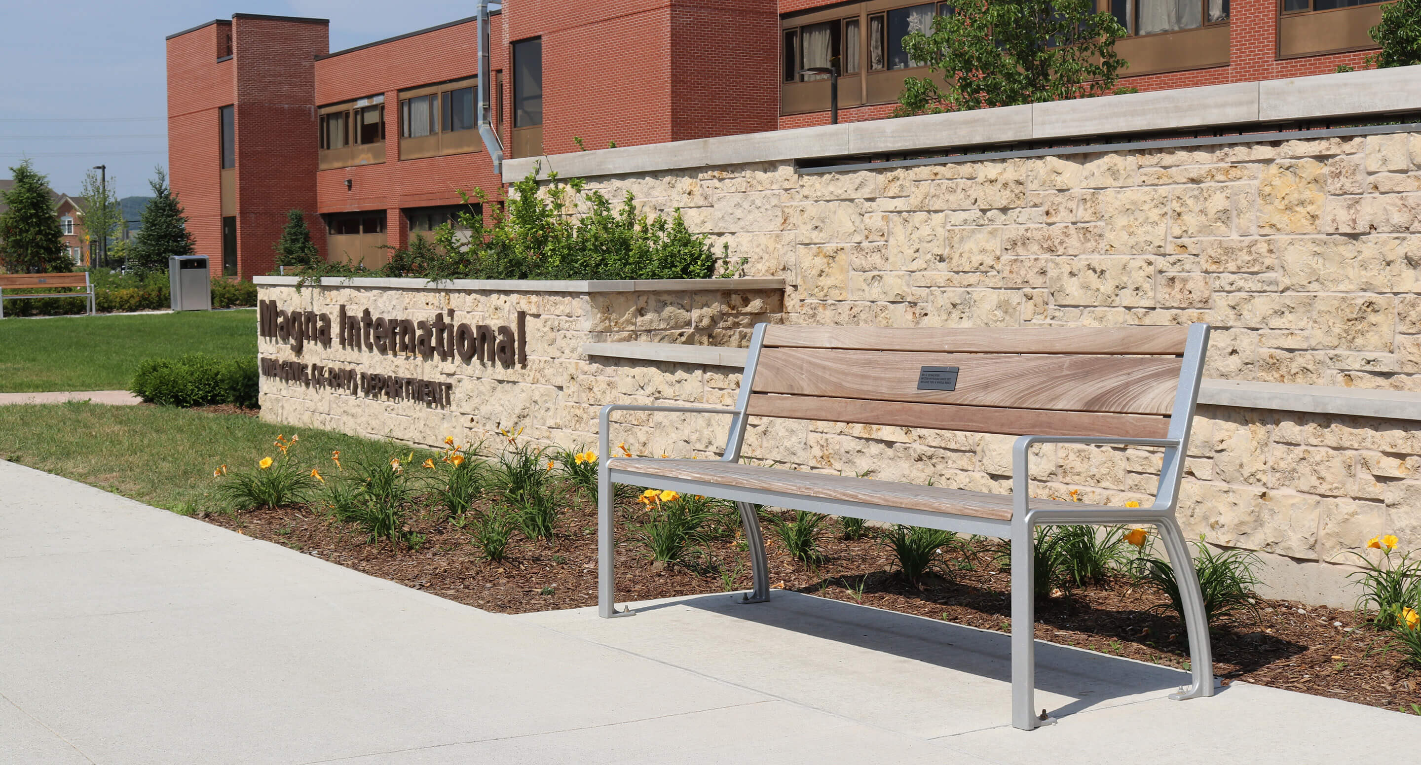 milton-district-hospital-970-bench-plaque Un banc en bois posé sur une allée en béton, devant un mur en pierre sur lequel figure l'enseigne « Magna International ».