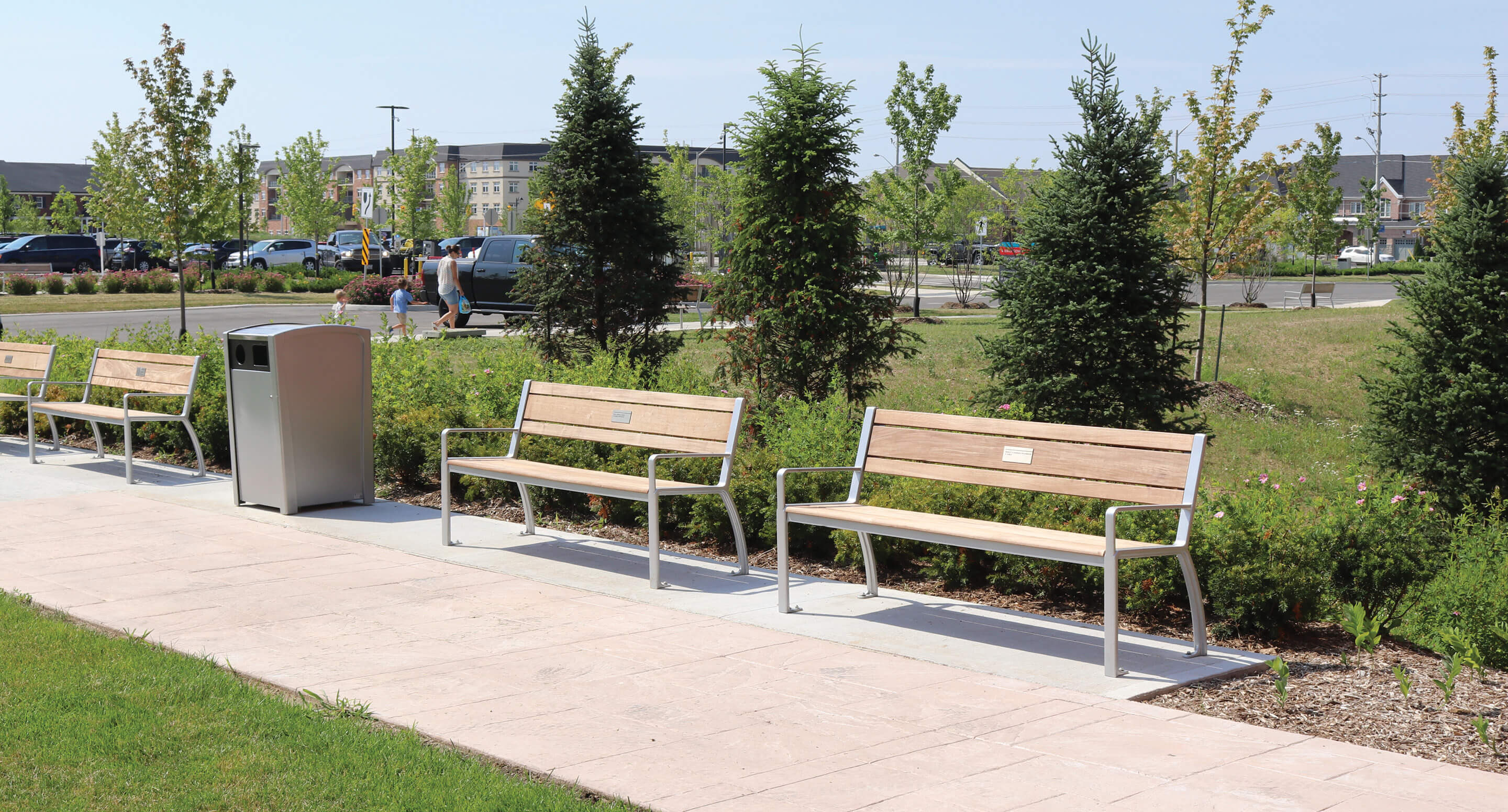 milton-district-hospital-970-benches-plaque-waste-container-parking-lot Trois bancs en bois avec une structure métallique, disposés le long d'une allée en béton dans un parc.