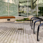 Une cour pavée avec des râteliers à vélos noirs, un banc en bois posé sur un socle en béton, et des baies vitrées ornées de motifs de poissons et de visages en vinyle blanc.