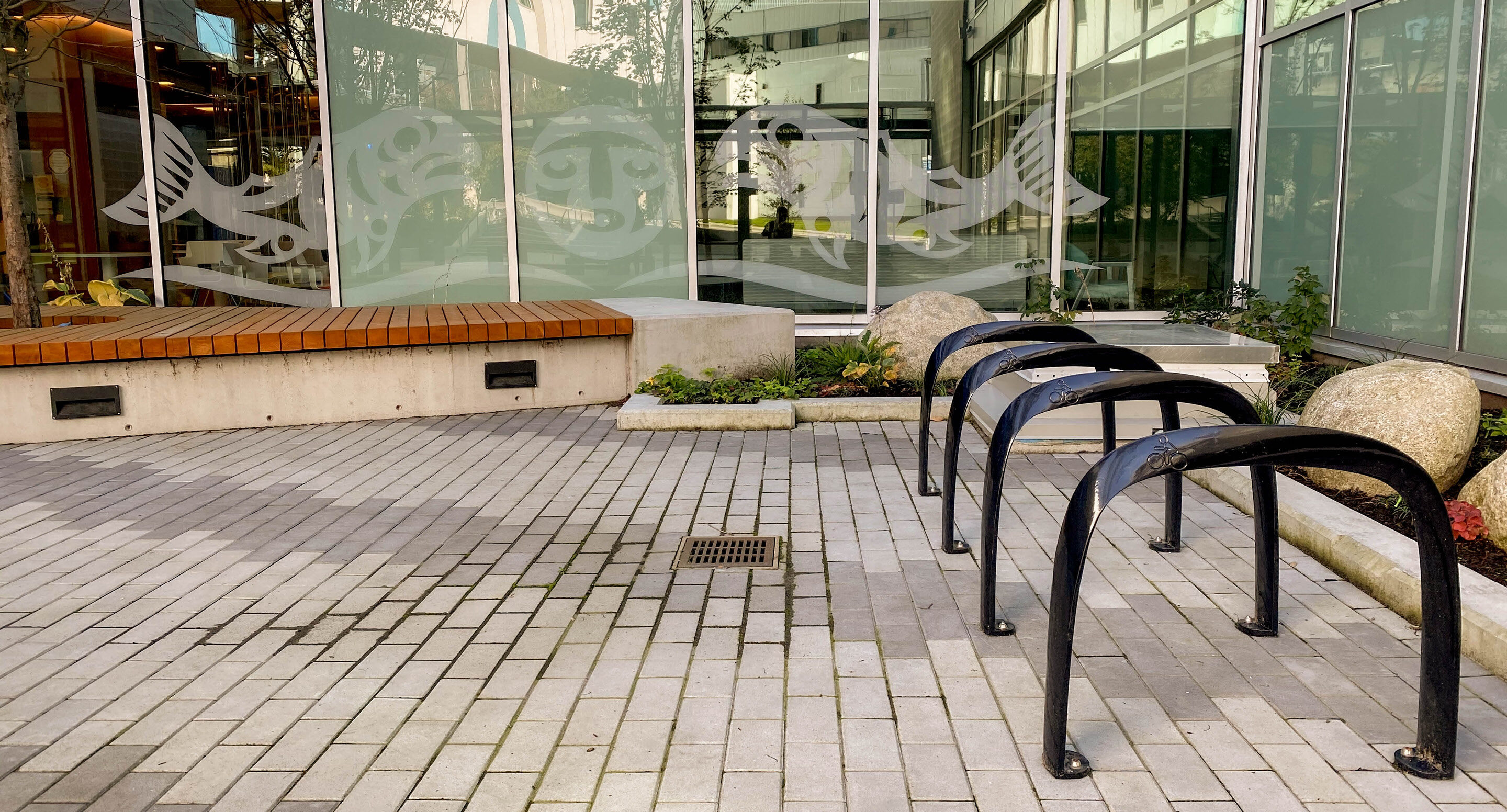 royal-columbian-hospital-720-bike-racks Une cour pavée avec des râteliers à vélos noirs, un banc en bois posé sur un socle en béton, et des baies vitrées ornées de motifs de poissons et de visages en vinyle blanc.