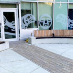Two curved wooden benches with concrete bases sit on a paved patio outside the Royal Columbian Hospital Substance Use Wellness Centre, in front of windows featuring salmon-themed graphic decals.