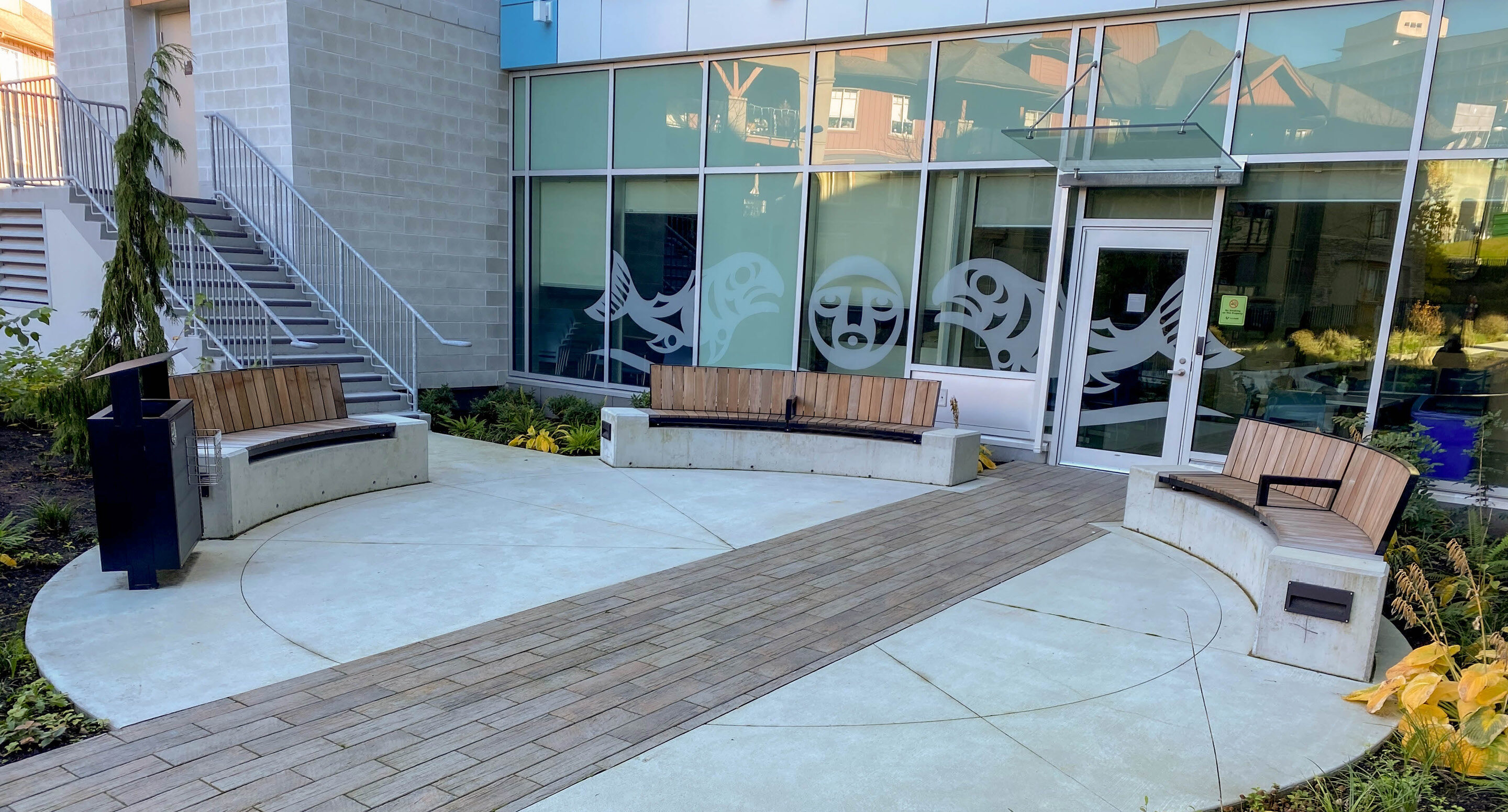 royal-columbian-hospital-ogden-bench-entrance-2 Trois bancs incurvés en bois et en béton disposés sur une place à l'extérieur d'un bâtiment aux baies vitrées ornées d'œuvres d'art d'inspiration autochtone.