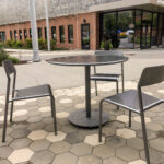 Trois chaises en métal disposées autour d'une table ronde, sur une terrasse pavée hexagonale située à l'entrée d'un immeuble portant l'inscription « Brookline Place Parking ».