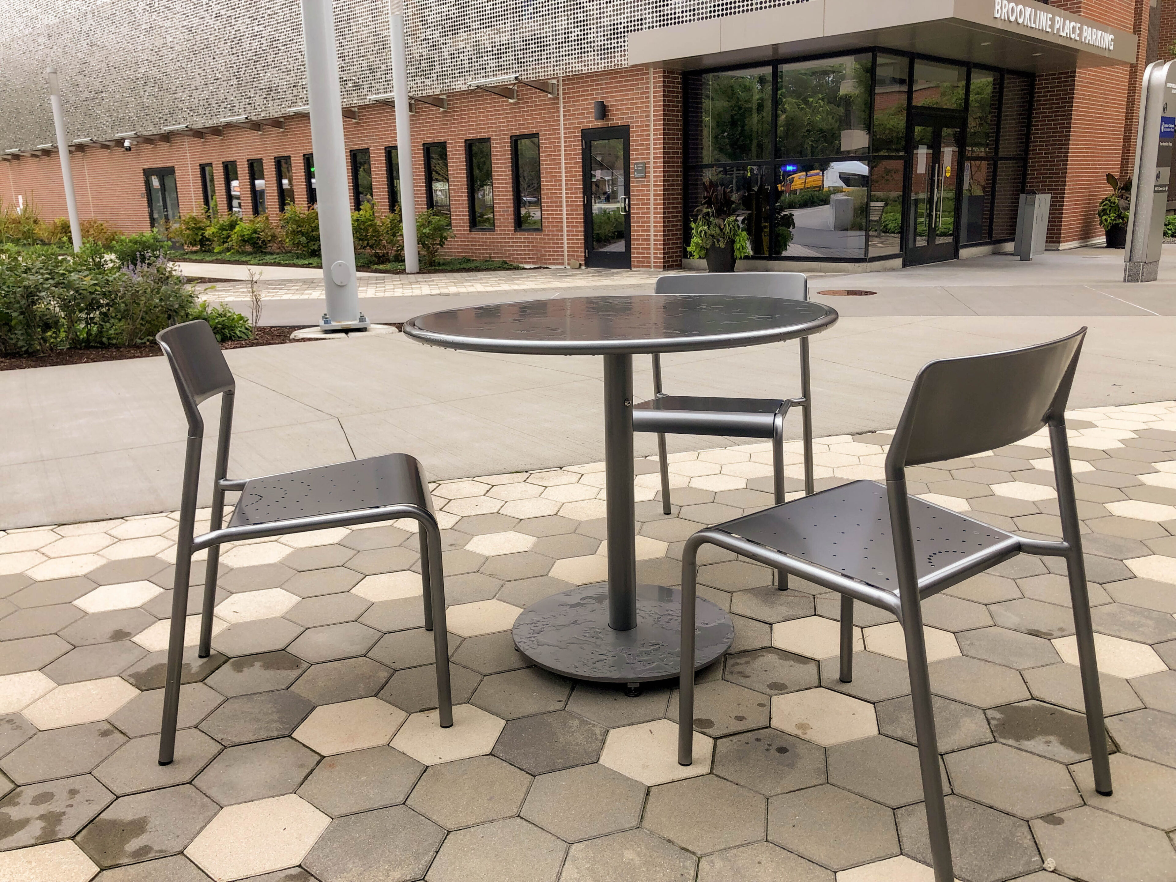 two-brookline-boston-childrens-foro-tables-chairs-3 Trois chaises en métal disposées autour d'une table ronde, sur une terrasse pavée hexagonale située à l'entrée d'un immeuble portant l'inscription « Brookline Place Parking ».