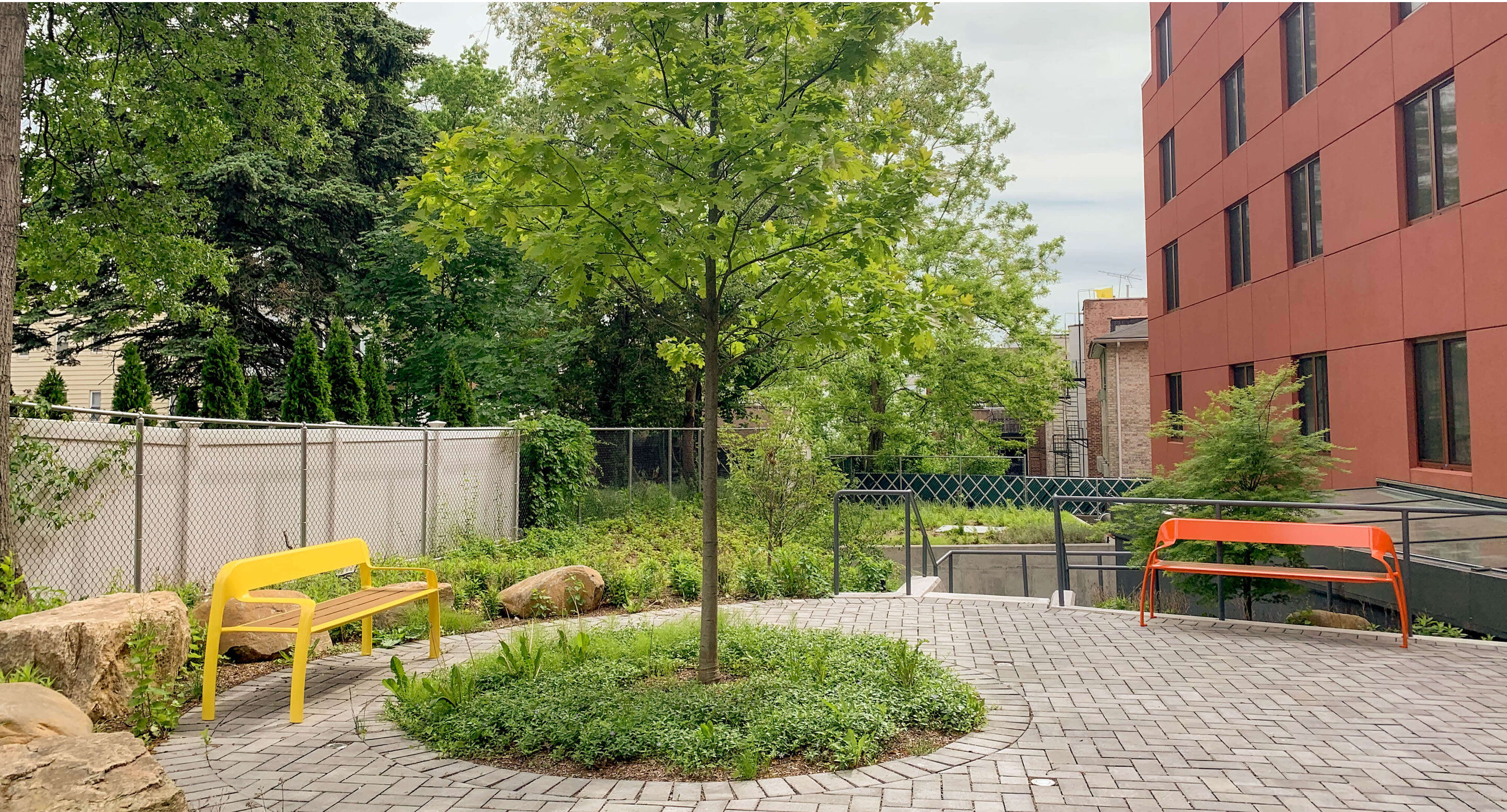 woodlawn-senior-living-custom-benches Une terrasse pavée avec un arbre au centre, un banc jaune, un banc orange et un bâtiment rouge.
