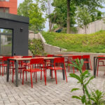 Une terrasse extérieure avec des chaises rouges et des tables en bois, aménagée sur une terrasse pavée à côté d'un immeuble moderne.