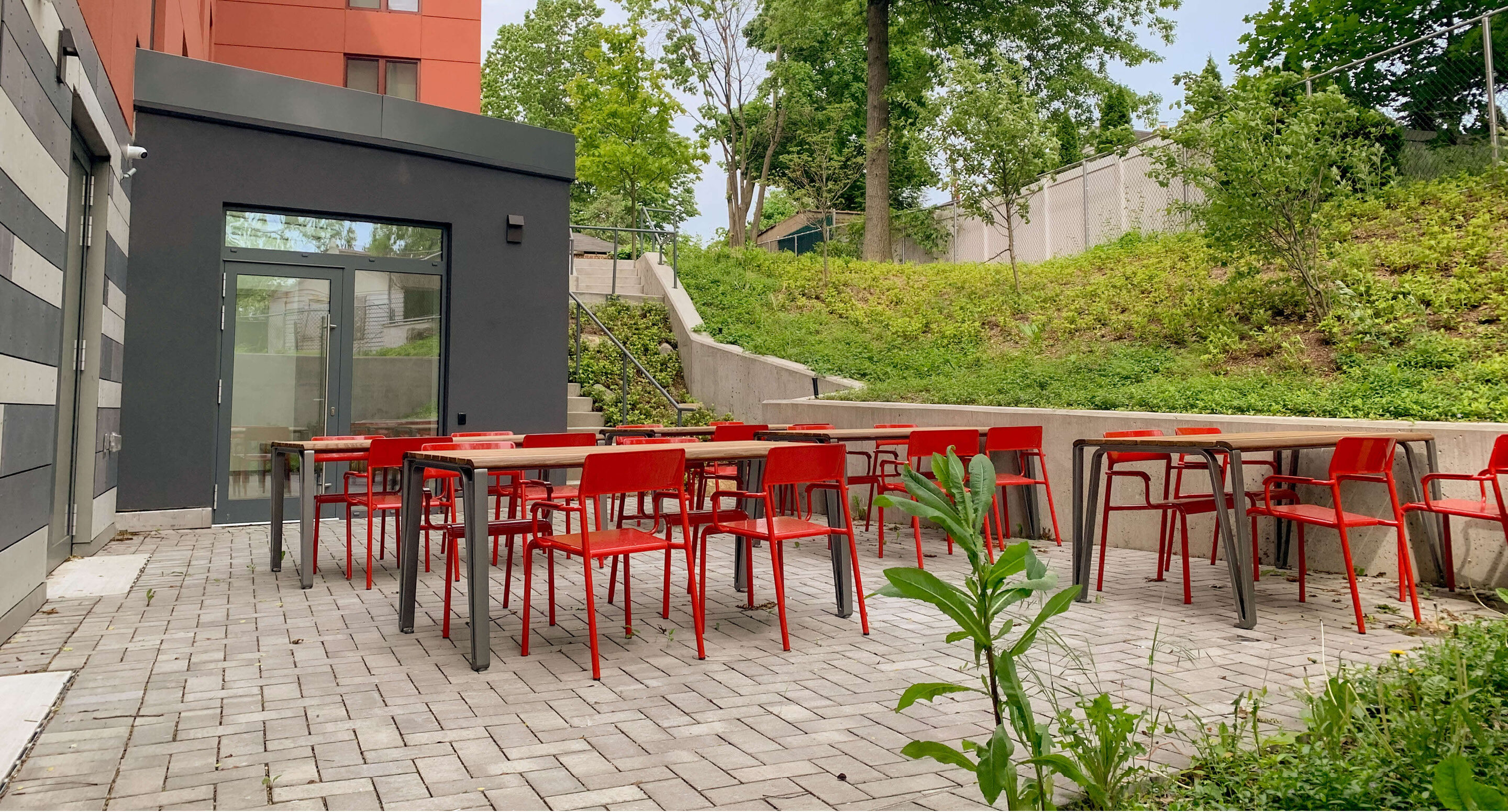 Une terrasse extérieure avec des chaises rouges et des tables en bois, aménagée sur une terrasse pavée à côté d'un immeuble moderne.