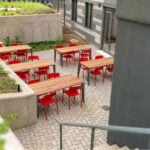 An outdoor patio at Woodlawn Senior Living with wooden tables and red chairs, located in a sunken area next to a concrete retaining wall with greenery.