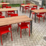 Terrasse extérieure avec des tables en bois et des chaises en métal rouge, sur un sol pavé.