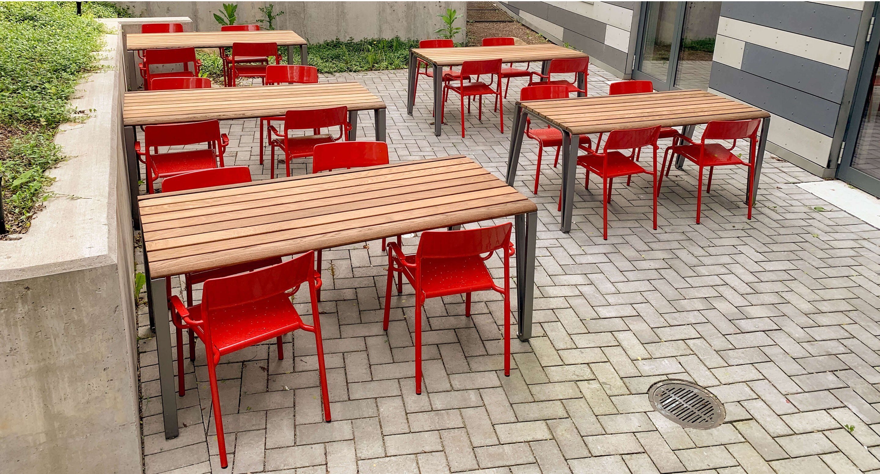 Terrasse extérieure avec des tables en bois et des chaises en métal rouge, sur un sol pavé.