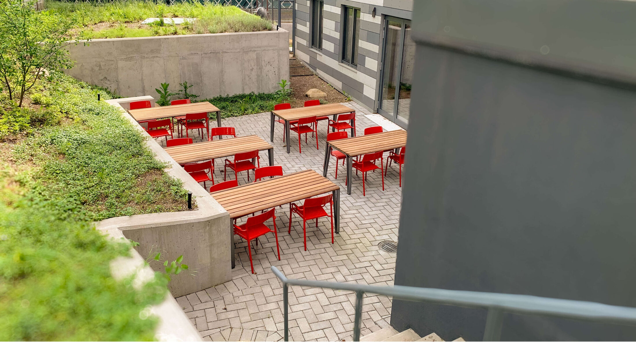 An outdoor patio at Woodlawn Senior Living with wooden tables and red chairs, located in a sunken area next to a concrete retaining wall with greenery.